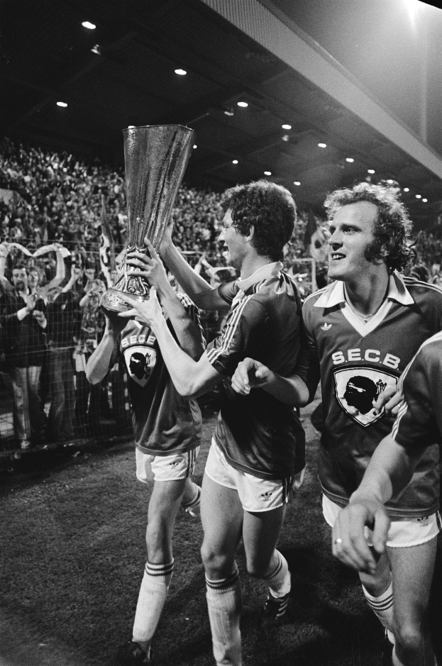 Eine Gruppe von Männern in Schwarz-Weiß-Kleidung hält einen Pokal auf einem Fußballfeld hoch, feiert ihren Sieg im FA-Cup-Finale, umgeben von einer Menge hinter einem Zaun und beleuchtet von Deckenlampen.