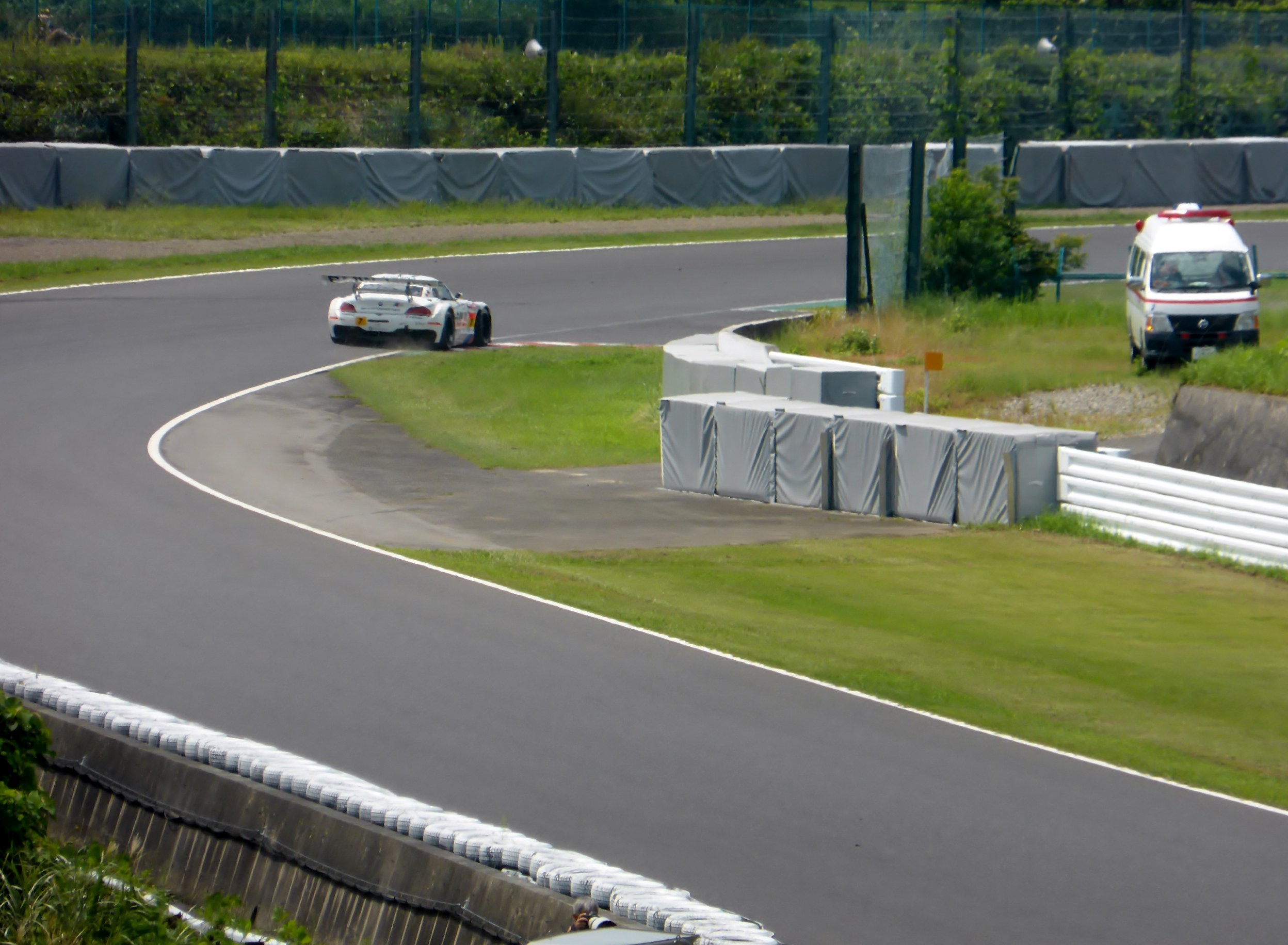 Ein Rennwagen, der auf einer Rennstrecke fährt, mit einem Polizeiwagen im Hintergrund, umgeben von Gras, Pflanzen, Bäumen und einem Zaun, mit einem Fahrzeug auf der rechten Seite und Text unten.