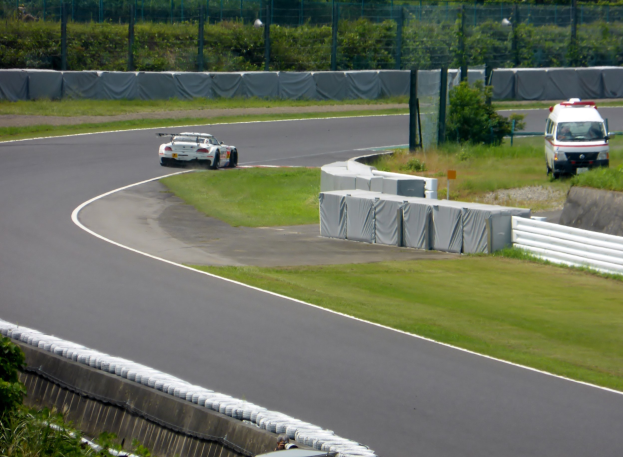 Ein Rennwagen, der auf einer Rennstrecke fährt, mit einem Polizeiwagen im Hintergrund, umgeben von Gras, Pflanzen, Bäumen und einem Zaun, mit einem Fahrzeug auf der rechten Seite und Text unten.