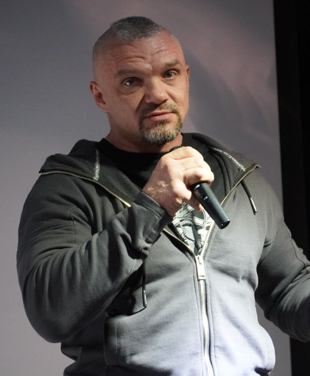 Ein Mann in einer schwarzen Kapuzenjacke und einem schwarzen Anzug mit einem weißen Hemd steht selbstbewusst vor einer Wand und hält ein Mikrofon in der Hand und sieht aus wie ein Komiker.