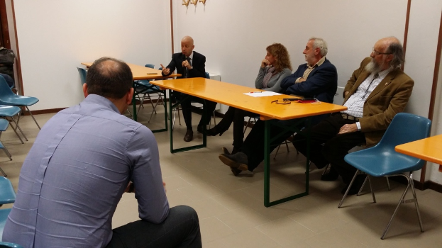 Eine Gruppe von Menschen sitzt um einen Tisch in einem Raum, wobei ein Mann in einer schwarzen Blazer ein Mikrofon in der Mitte hält.