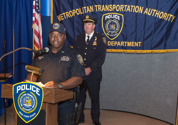 Zwei Polizeibeamte stehen vor einem Rednerpult, einer hölt Papiere und spricht in ein Mikrofon, mit einer Fahne, einem Tisch und einer Tafel in der Nähe, und einer Tafel mit Symbolen und Text im Hintergrund.
