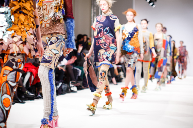 Eine Gruppe Von Frauen Geht Den Laufsteg Bei Der London Fashion Week entlang, Mit Einer Zuschauerschaft Im Hintergrund Unter Hellen Lichtern.