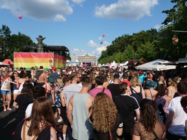 Eine große Menschenmenge geht eine Straße mit Zelten, Bäumen, Polen, Lichtern und einer Statue entlang, im Hintergrund Gebäude und ein Himmel mit Wolken und Ballons, während des Berliner Christopher Street Day.