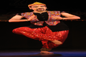 Ein Mann in roter und schwarzer Kleidung tanzt auf der Bühne mit dunklem Hintergrund, die Augen geschlossen und der Mund leicht geöffnet.