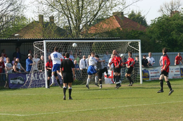 Fußballspieler spielen auf einem Feld mit einem Tor hinter ihnen, während Zuschauer zuschauen; Bäume und Häuser sind im Hintergrund zu sehen.