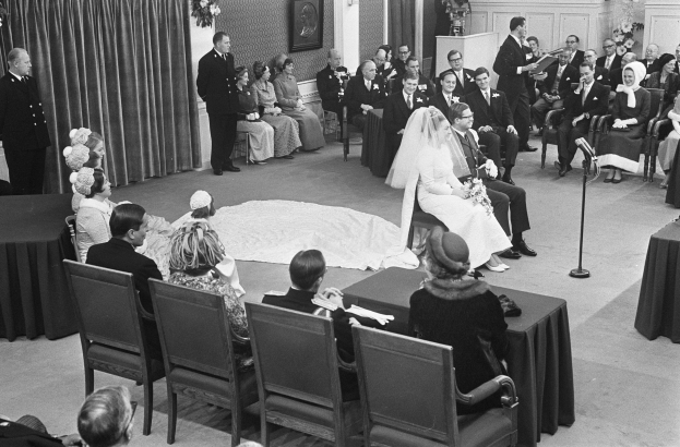 Schwarzes und weißes Foto einer Hochzeitszeremonie mit sitzenden und stehenden Menschen, einem Mann, der in ein Mikrofon spricht, und einer Frau, die einen Strauß hält.