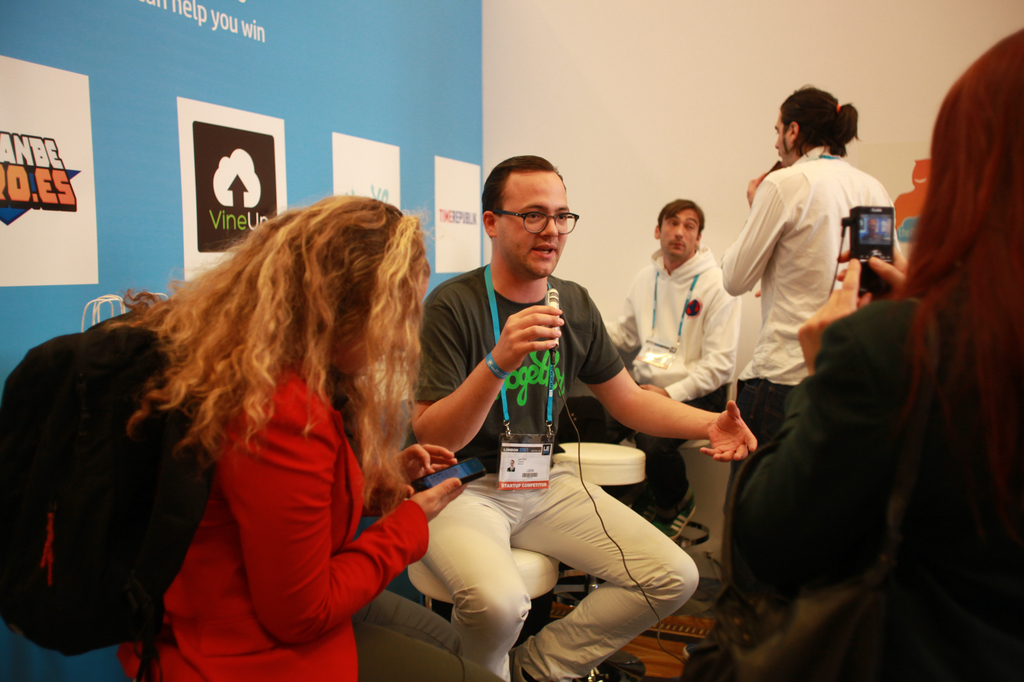 Ein Interview findet statt mit Menschen, die Mikrofone und eine Kamera halten, mit Personen im Vordergrund und einem Banner links und einem klaren Hintergrund.