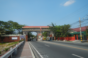 Eine Straße mit Fahrzeugen, ein Fußweg mit Geländern auf der linken Seite, Bäume und Gebäude auf beiden Seiten der Straße, ein Tor mit einer Namensschild, Strommasten mit Drähten und ein bewölkter Himmel im Hintergrund.