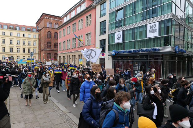 Eine große Gruppe von Menschen geht eine Straße in Stockholm, Schweden, entlang, einige tragen Masken und Taschen, andere halten Schilder, während eine Person links filmt, Gebäude, Schilder, und ein klarer blauer Himmel im Hintergrund.