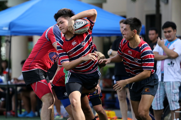 Eine Gruppe von Menschen spielt ein Spiel im Freien mit einem Jungen in der Mitte, der einen Ball hält, während andere stehen und im Hintergrund bei einem blauen Zelt zuschauen.