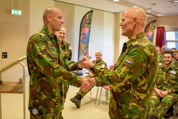 Zwei Männer in Militäruniformen geben sich die Hand vor einer sitzenden Gruppe, einem Tisch, einem Geländer, Bannern, einer Tür, einem Schild und einer Deckenbeleuchtung im Hintergrund.