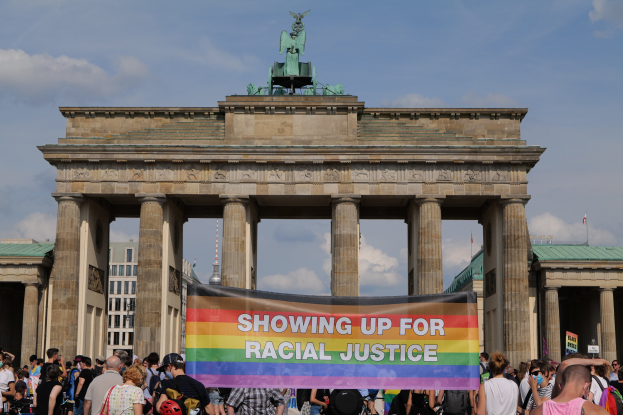 Eine Gruppe von Menschen steht vor dem Brandenburger Tor in Berlin, Deutschland, mit einer "Racial Justice"-Schleife, wobei die Säulen und die Statue des Tors sowie Gebäude und ein bewölkter Himmel im Hintergrund zu sehen sind.