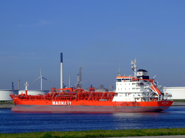 Ein großes rotes und weißes Öltankschiff schwimmt auf einem Gewässer, umgeben von Gras und einer Straße, mit Windrädern und einem klaren blauen Himmel im Hintergrund.