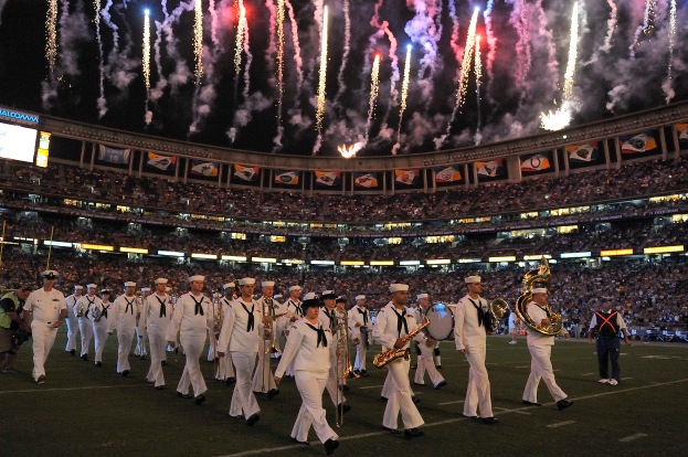Eine Gruppe von Menschen in weißen Uniformen marschiert auf einem Fußballfeld, einige halten Musikinstrumente, mit einer großen Menge im Stadion und Bannern im Hintergrund, Bildschirme links und Feuerwerk am Himmel.