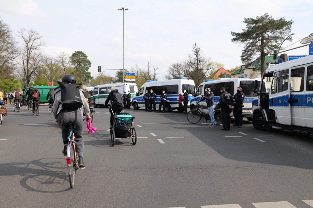 Eine Gruppe von Menschen, die auf Fahrrädern eine Straße entlangfahren, mit Polizeiwagen und anderen Fahrzeugen, einige tragen Helme und haben Taschen, andere schieben Kinderwagen, mit Bäumen, Laternenmasten, Verkehrsampeln, Verkehrsschildern, Gebäuden und einem bewölktem Himmel im Hintergrund.
