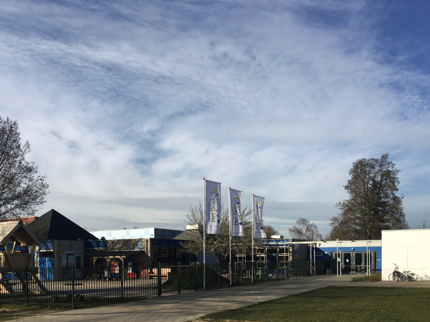 Gro├če Geb├Ąude mit einem grasigen Bereich davor, umgeben von einem Metallzaun, B├Ąumen, Bannern, einem geparkten Fahrrad, n├Ąheren H├Ąusern und einem bew├Âlktem Himmel, identifiziert als der neue Spielplatz der Schule.