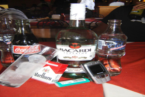 Alkoholflasche, Wasserflaschen, eine Coca-Cola-Flasche, ein Glas mit Eiswürfeln, ein Feuerzeug und ein Mobiltelefon auf einem Tisch mit Stühlen und Menschen, die Wein trinken.