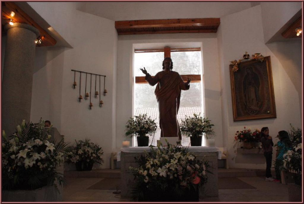 Ein Innenblick eines Gebäudes mit Kindern, die auf dem Boden stehen, einer Skulptur vor einer mit Stoff bedeckten Wand, Topfpflanzen, einem Fenster, einer Säule mit Holz basis und Lampen und einem an der Wand angebrachten Photo frame.