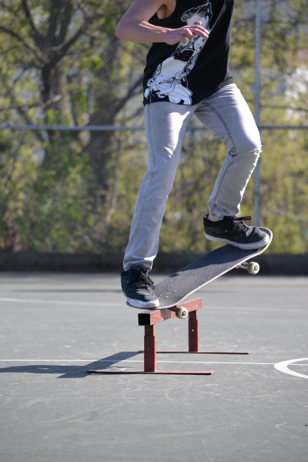 Eine Person in einem T-Shirt fährt Skateboard Outdoor mit einem Metallobjekt auf dem Boden, mit einem Netz, Metallstangen, Pflanzen und Bäumen im Hintergrund.