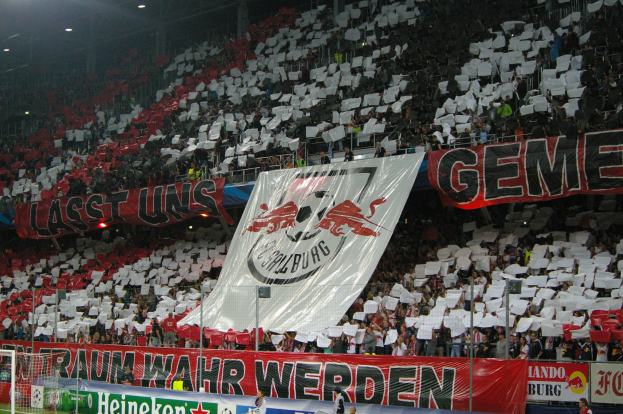 Ein überfülltes Fußballstadion mit sitzenden und stehenden Zuschauern, Banner mit Text und Logos, eine sichtbare Torstange und Überdachung.