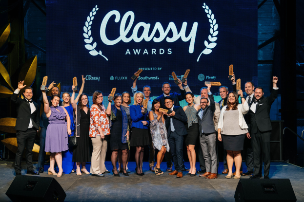 Eine Gruppe von Menschen auf einer Bühne, die strahlen und Auszeichnungen halten, mit Lautsprechern unten und einem Hintergrundbild "Classy Awards 2019".