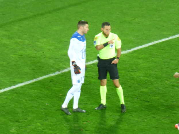 Ein Schiedsrichter in gelbem T-Shirt und schwarzen Shorts spricht mit einem Füßballer in weißer und blauer Uniform auf dem Feld, beide tragen Turnschuhe.