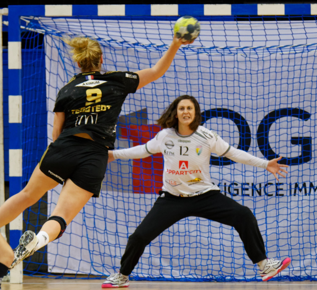 Zwei Frauen beim Handballspielen in der Nähe eines Tors, eine hält den Ball, mit einem Netz und einem Schild im Hintergrund.