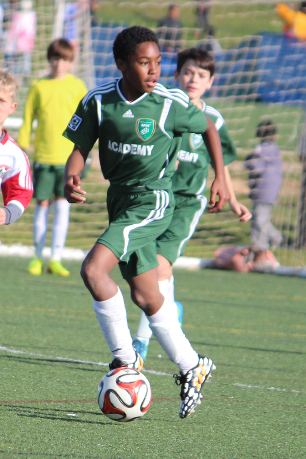 Zwei Personen spielen Fußball in einem Stadion mit einigen Zuschauern und einem Netz im Hintergrund.
