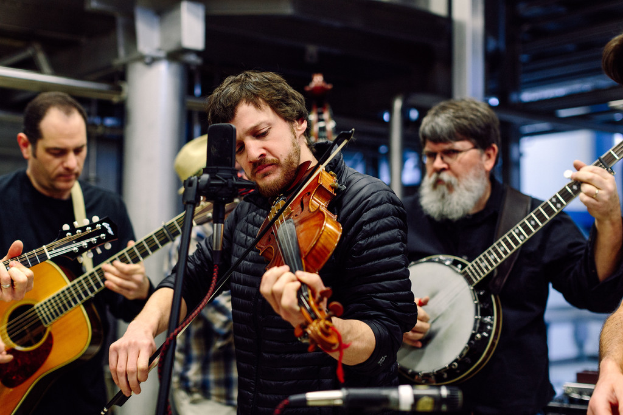 Eine Gruppe von Musikern in schwarzer Kleidung, die verschiedene Instrumente spielen, mit einem Mikrofon vor dem Geigenvirtuosen und einem Gebäude im Hintergrund.