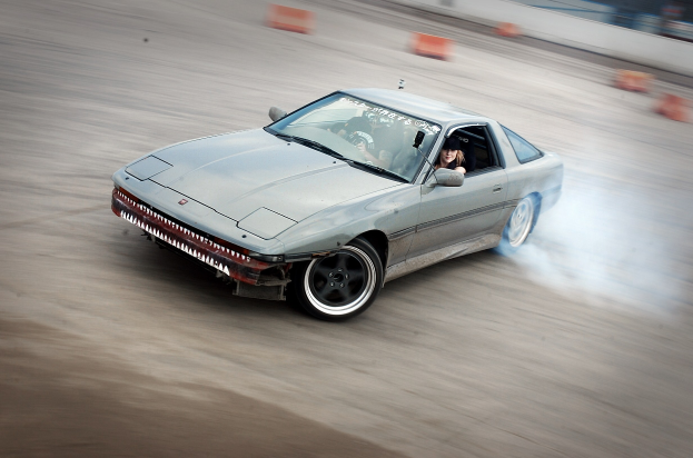 Eine Person mit Helm fährt ein Auto auf einer Rennstrecke, mit Rauch, umgeben von orangefarbenen Gegenständen, mit einem unscharfen Hintergrund, der hohe Geschwindigkeit andeutet.