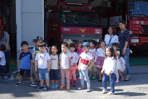 Eine Gruppe von Kindern steht vor einem Löschwagen auf einem Feuerwehrareal, einige tragen Mützen, mit zusätzlichen Löschwagen im Hintergrund.