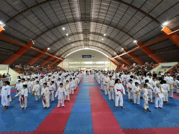 Große Gruppe von Menschen auf einer blauen und roten Matte in einer Turnhalle mit anderen auf der Treppe sitzend, Deckenlampen und Hintergrundtafeln, die auf ein Taekwondo-Event hinweisen.