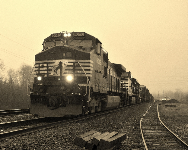 Ein Zug auf Bahnschienen mit Autos, Strommasten, Bäumen und einem Himmel im Hintergrund.