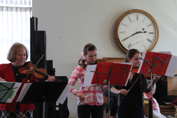 Drei Frauen auf der Bühne mit Geigen stehend und Musikständer vor sich, während ein sitzendes Publikum zuhört; eine Uhr ist an der weißen Wand im Hintergrund angebracht, mit einem weißen Vorhang in der linken Ecke.
