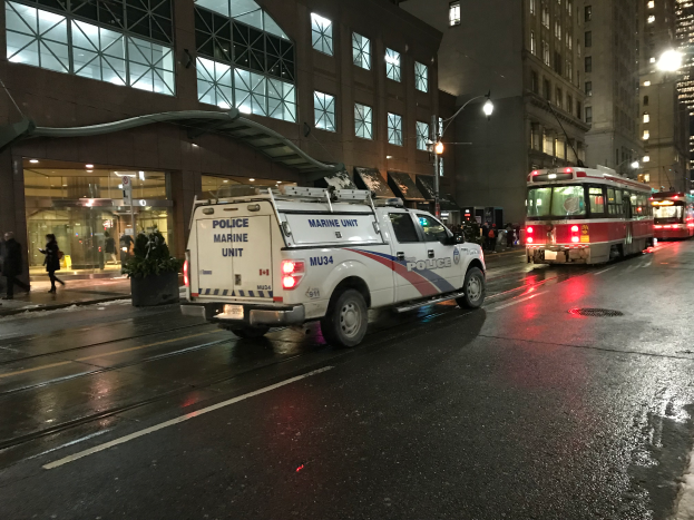 Ein Polizeiwagen parkt nachts am Straßenrand einer Stadt, mit Fahrzeugen auf der Straße, Fußgängern auf dem Gehweg, Topfpflanzen, Straßenlaternen, Gebäuden mit Fenstern und einem bewölkten Himmel.