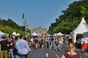 Eine Menschenmenge, die eine Straße entlanggeht, die von Zelten, Fahrzeugen und Bäumen gesäumt ist, mit einem Bogen und einem klaren blauen Himmel im Hintergrund und Polen mit Fahnen auf der linken Seite, die wahrscheinlich das Oktoberfest in München, Deutschland, darstellen.