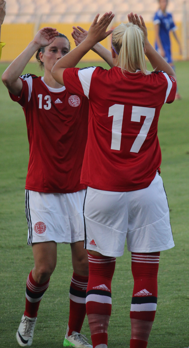 Zwei Frauen in roten T-Shirts und weißen Shorts stehen auf einem Fußballfeld und klatschen in die Hände, während im Hintergrund eine Person in einem blauen Kleid, eine gelbe Tafel und ein paar Stühle zu sehen sind.