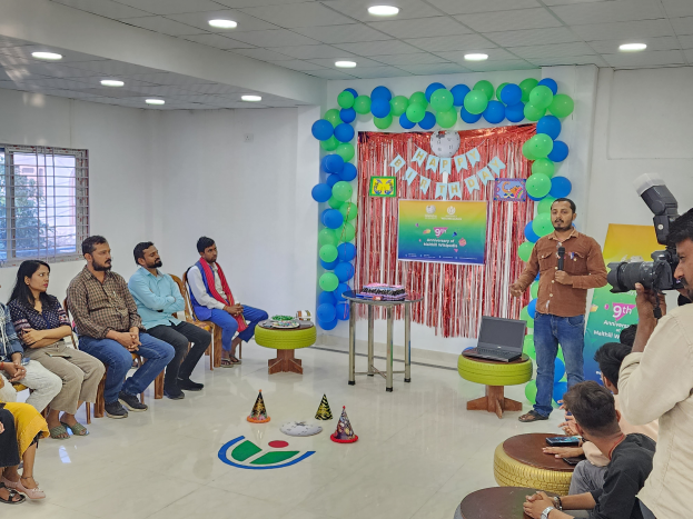 Eine Gruppe von Menschen sitzt auf Stühlen vor einer Menge, ein Mensch hält ein Mikrofon, ein Laptop auf einem Hocker, ein Tisch mit verschiedenen Gegenständen, eine Fahne mit Text, Luftballons, ein Vorhang, ein Fenster und eine Decke mit Deckenleuchten, feiern einen 9. Geburtstag.