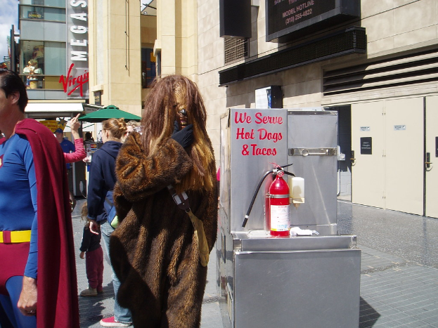 Eine Menschenmenge, darunter einige in Kostümen, versammelte sich vor einem Gebäude mit sichtbarer Beschriftung und Türen, mit einer Kiste und einem Feuerlöscher in der Nähe.