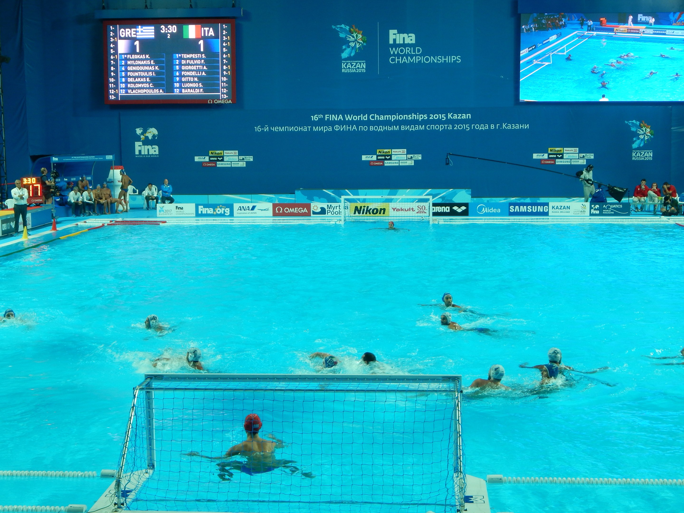 Eine Gruppe von Menschen spielt Wasserball in einem Schwimmbad während der FINA-Weltmeisterschaften 2016 in Kazan, mit Zuschauern auf der linken Seite und einem Bildschirm mit Event-Branding im Hintergrund.
