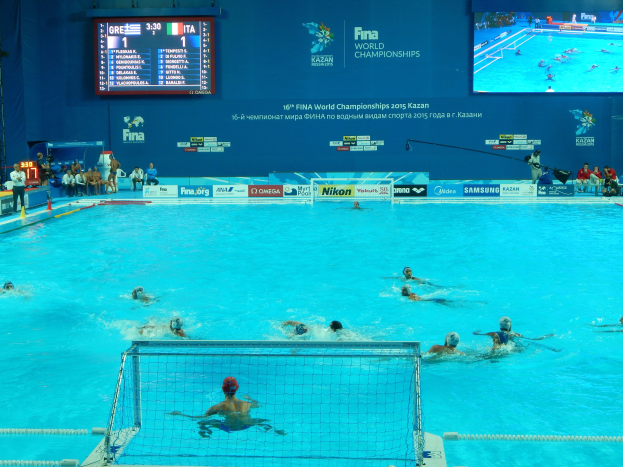 Eine Gruppe von Menschen spielt Wasserball in einem Schwimmbad während der FINA-Weltmeisterschaften 2016 in Kazan, mit Zuschauern auf der linken Seite und einem Bildschirm mit Event-Branding im Hintergrund.