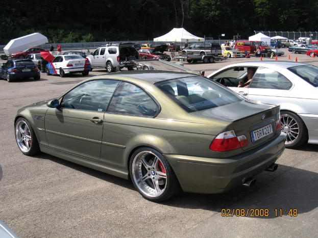 Eine Außenaufnahme mit mehreren Fahrzeugen auf einem Parkplatz, einem weißen Zelt in der Ferne, zahlreichen Bäumen, einigen Menschen und einem roten Schirm sowie zwei Autos im Vordergrund.