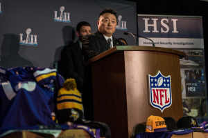 Ein Mann in einem schwarzen Blazer, einem wei├čem Hemd und einer schwarzen Krawatte spricht an einem Podium mit einem Mikrofon und Logo, vor einer Menge. Blaue und gelbe Merchandise und ein "Super Bowl LII"-Banner sind im Hintergrund zu sehen.