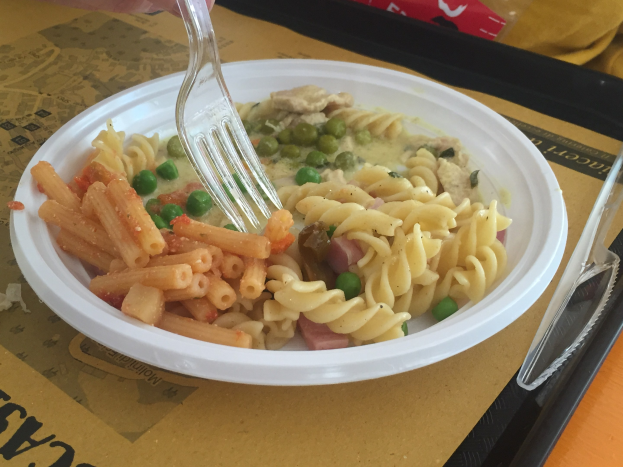 Eine Person hält eine Gabel über eine Schüssel mit Nudeln, Erbsen und Schinken, mit einem Tablett mit einem Löffel und einer Abdeckung auf dem Tisch.