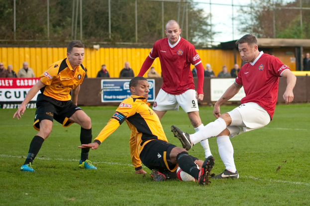 Spieler in blauen und roten Uniformen spielen ein Spiel auf einem Rasenfeld mit einem Ball, während Zuschauer außerhalb des Feldes stehen und sie anfeuern, mit einem Baum und Himmel im Hintergrund.