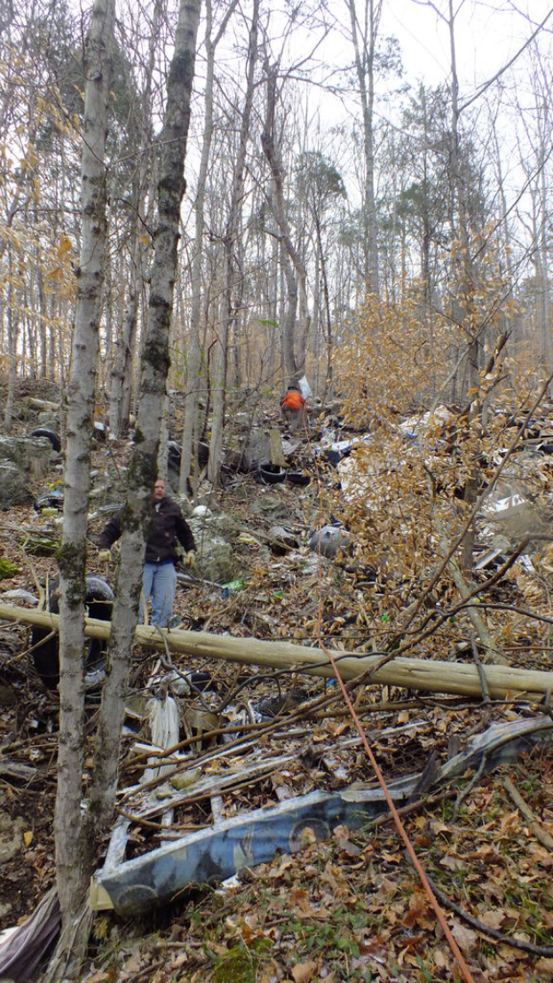 Ein Mann in einer schwarzen Jacke posiert vor einem Waldpanorama mit trockenen Bäumen im Vordergrund und hohen Bäumen im Hintergrund.