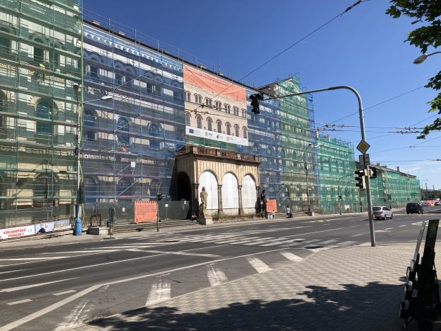 Großer mehrstöckiger Bau im Bau in Berlin, umgeben von Straßeninfrastruktur, Fahrzeugen, Fußgängern und Bäumen unter einem bewölkten Himmel.
