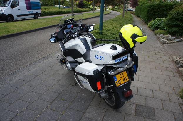 Ein polizeiliches Motorrad steht am Straßenrand mit einem Helm darauf, umgeben von Bäumen, Pfählen, Gebäuden, Pflanzen, Gras, Steinen und anderen Fahrzeugen.