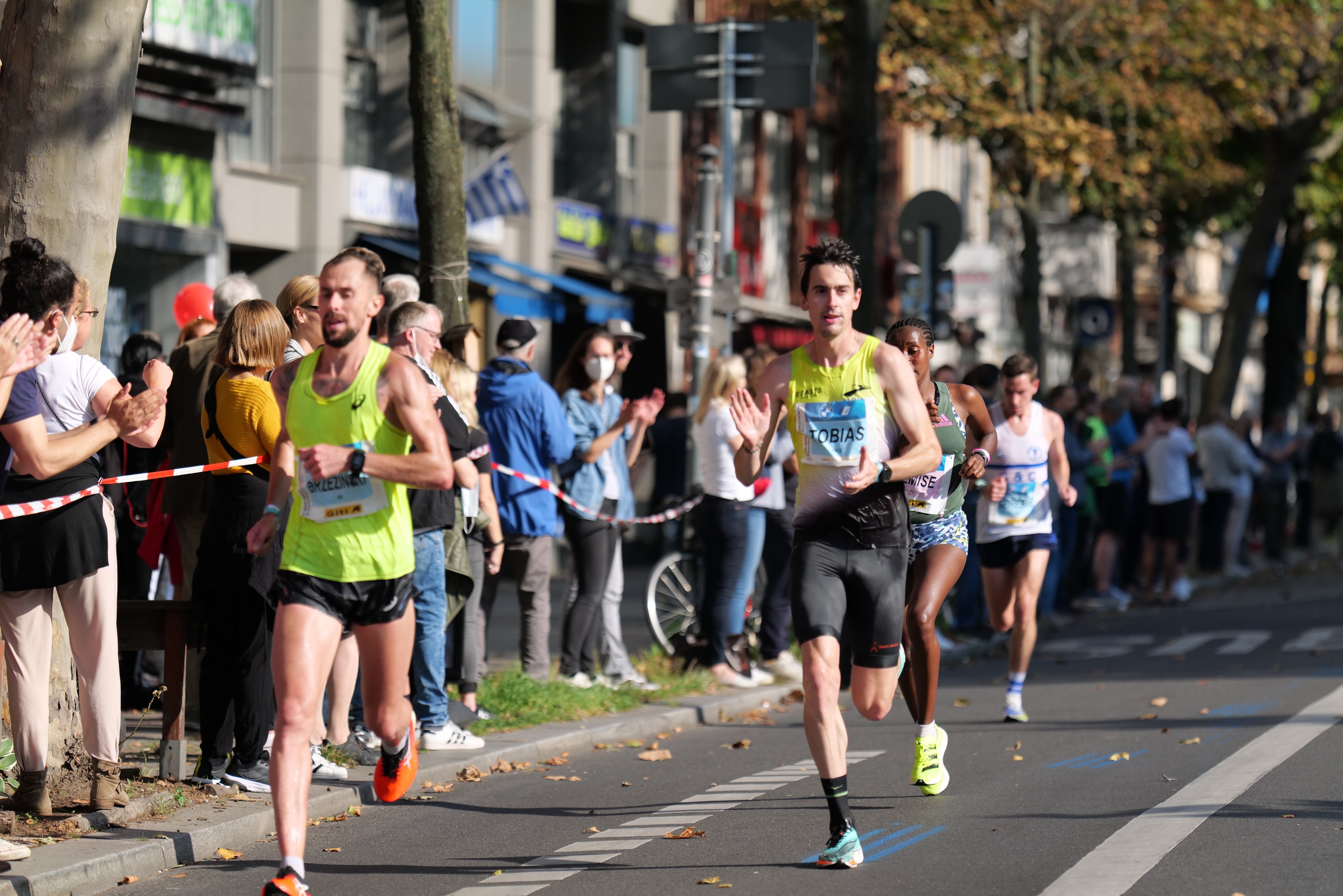 Gruppe von Läufern bei einem Marathon auf einer Stadtstraße mit Zuschauern auf der linken Seite, Bäumen, Gebäuden und einem klaren blauen Himmel im Hintergrund.